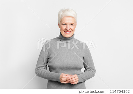 Cheerful elderly European woman with clasped hands, illustrating s3niorlife contentment on grey background 114704362