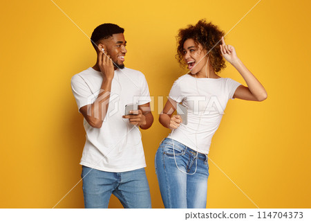 Joyful millennial black man and woman in earphones listening to music on cellphones and smiling to each other, orange background Joyful millennial black man and woman in earphones listening to music on cellphones and smiling to each other, orange background 114704373