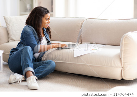 Busy African American woman is sitting comfortably on the floor, focused on using her laptop. She appears to be typing intently, possibly working or studying. 114704474