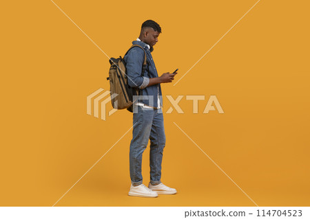 African american guy projects a multiethnic vibe while casually checking his phone, isolated on orange background African american guy projects a multiethnic vibe while casually checking his phone, isolated on orange background 114704523