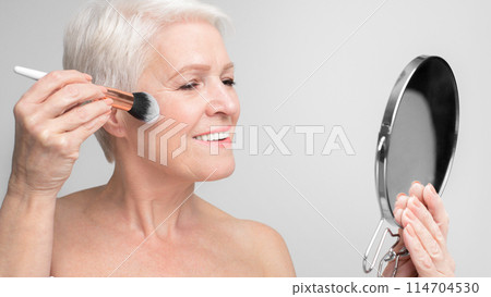 An elegant elderly european woman applies blush with a brush while looking into a mirror, representing s3niorlife and self-care 114704530