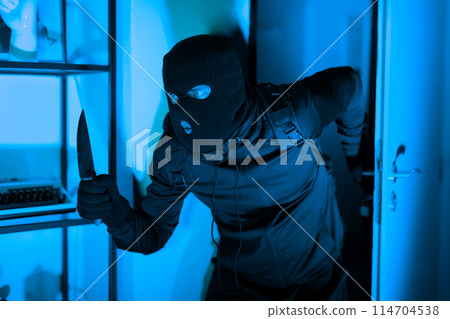 A masked burglar in black clothing holding a knife is caught in the act during a nighttime home invasion, illuminated by eerie blue light 114704538