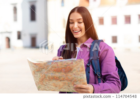 A cheerful millennial lady in purple outfits stands as a quintessential student outdoors while studying a map in a European city 114704548