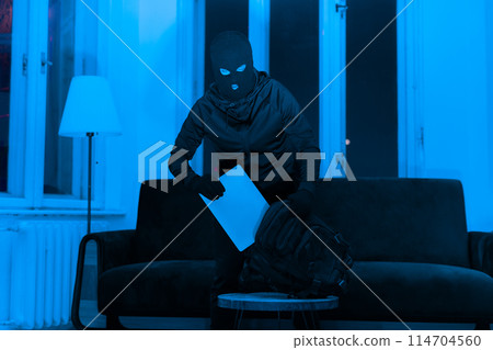 A burglar appears to be stealing laptop, rifling through a backpack in an apartment at night, creating an unsettling atmosphere 114704560
