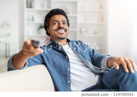 A cheerful black man sits comfortably on a sofa holding a TV remote, likely changing channels or about to watch television 114704591