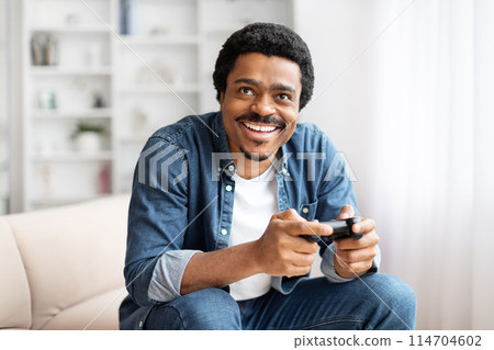 An african american man displays a broad smile while holding a game controller, enjoying gaming at home 114704602