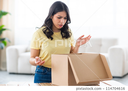A muslim woman at home showcasing a disappointed expression as she inspects an open cardboard box parcel A muslim woman at home showcasing a disappointed expression as she inspects an open cardboard box parcel 114704616