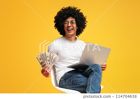 Online job. Excited black guy working on laptop and showing bunch of money, orange panorama background with free space Online job. Excited black guy working on laptop and showing bunch of money, orange panorama background with free space 114704638