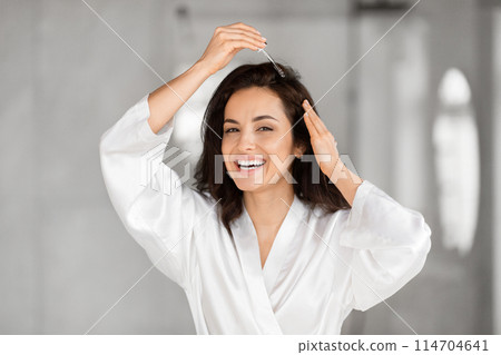 A joyful woman in a crisp white bathrobe playfully touches her hair in a bright, airy room, her expression exuding happiness and relaxation 114704641