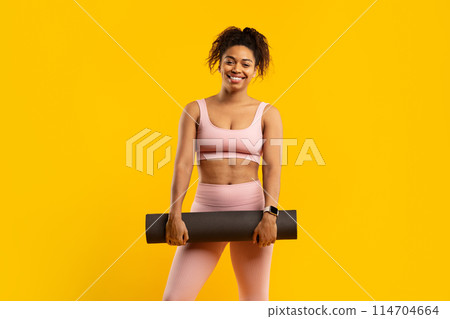 Smiling young african american woman in sportswear holding a yoga mat, set against a vibrant yellow background 114704664