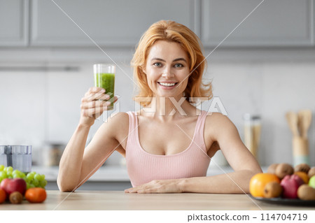 A cheerful blonde woman presenting a healthy green smoothie in a modern kitchen with fruits in the background A cheerful blonde woman presenting a healthy green smoothie in a modern kitchen with fruits in the background 114704719