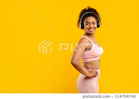 Fit black lady confidently poses with hands on hips, in workout clothing and headphones on yellow, copy space 114704748