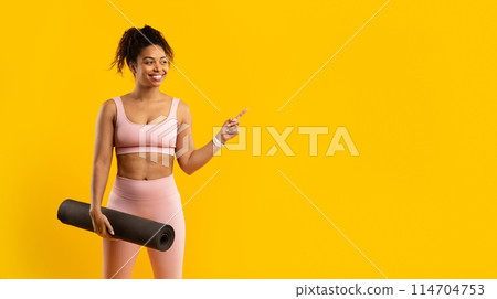 A cheerful african american woman in athletic wear holding a yoga mat and pointing to her side against a yellow background 114704753