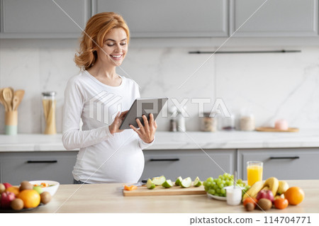 A european pregnant lady stands in a modern kitchen browsing a tablet, surrounded by fresh nutrition A european pregnant lady stands in a modern kitchen browsing a tablet, surrounded by fresh nutrition 114704771