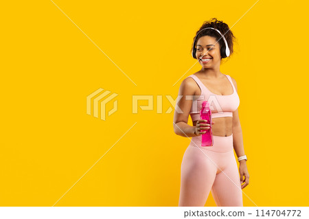 Joyful black lady with fitness outfit holding a water bottle, gazing off-camera on yellow background, copy space 114704772