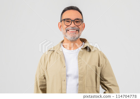 A cheerful senior man with a beard and glasses stands posing in a beige shirt against a gray background, exuding confidence and happiness 114704778