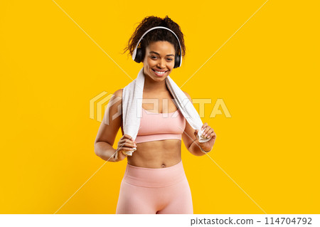 A joyful african american lady in fitness gear wearing headphones and a towel enjoys a workout break, isolated on yellow 114704792