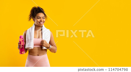 Radiant african american woman in fitness attire holding a pink water bottle, smiling with a towel around her neck on a yellow background Radiant african american woman in fitness attire holding a pink water bottle, smiling with a towel around her neck on a yellow background 114704798