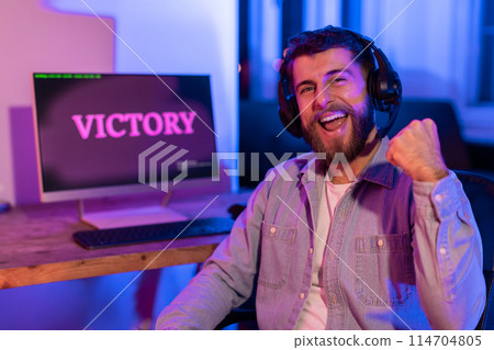 A passionate millennial guy fist pumps in front of a 'VICTORY' screen at home, illustrating the addictive excitement of gaming 114704805