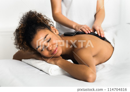 An african american lady revels in the comfort of a relaxing spa massage with hot stones carefully placed on her back by a masseuse. The image evokes a sense of warmth and care An african american lady revels in the comfort of a relaxing spa massage with hot stones carefully placed on her back by a masseuse. The image evokes a sense of warmth and care 114704816