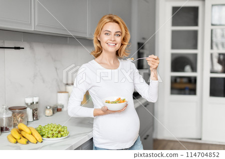 Cheerful european pregnant lady in a kitchen setting smiles while holding a nutritious bowl, emphasizing kitchen nutrition 114704852