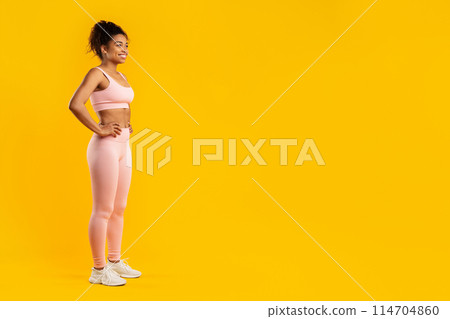 A toned african american woman poses in fitness wear with hands on her hips, isolated on a yellow background, copy space 114704860