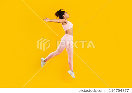 Dynamic image of an african american lady displaying energy and movement, mid-jump in pink workout clothing against a vibrant yellow backdrop Dynamic image of an african american lady displaying energy and movement, mid-jump in pink workout clothing against a vibrant yellow backdrop 114704877