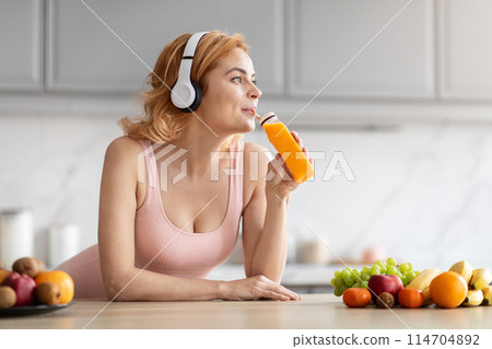 A young woman enjoys a glass of orange juice while wearing white headphones in a modern kitchen with fresh fruits 114704892