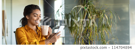 Cheerful african american lady manager chatting on cellphone, sitting with cup of coffee in modern office interior during break. Device for business remote, panorama with copy space 114704894