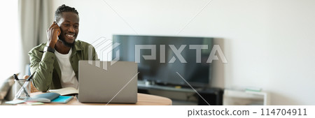 Remote career. Happy african american man talking on cellphone and using laptop, sitting at workplace at home. Freelancer guy having phone call and working on pc computer, copy space, banner 114704911