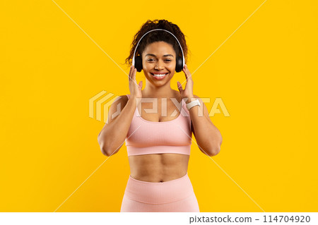A smiling young african american woman wearing a pink sports bra and leggings with headphones against a vibrant yellow background enjoys music 114704920