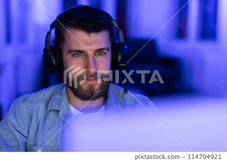 Calm man wearing a headset illuminated by calm blue light in front of a computer monitor, home interior, closeup Calm man wearing a headset illuminated by calm blue light in front of a computer monitor, home interior, closeup 114704921