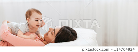 Motherhood concept. Loving young mom playing with her adorable child at home, relaxing on bed and bonding with cute infant kid, panorama with copy space 114704924