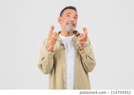 A hopeful old man with a beard is looking up, fingers crossed, wearing a beige shirt and white t-shirt on an isolated background Includes keywords like senior, elderly, old, man, isolated 114704932