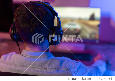 The guy gamer is deeply absorbed in his game, backlight illuminating his figure amidst the dark room's ambiance 114704951