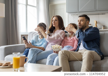 A european family shows diverse reactions of shock, curiosity, and amusement to a scene onscreen, embodying love and close relationships within a home environment 114704952