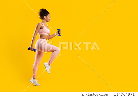 Determined african american lady engaged in a rigorous fitness routine with dumbbells, highlighting strength and focus, against an isolated yellow background 114704971