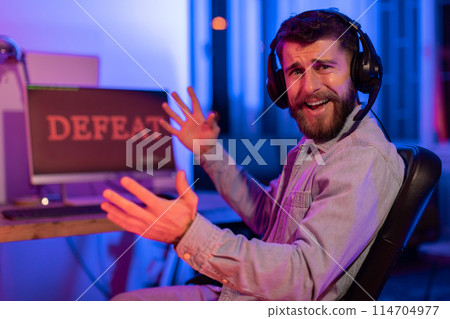 Perplexed gamer bearded guy reacts with frustration to a gaming defeat, displayed on his computer screen Perplexed gamer bearded guy reacts with frustration to a gaming defeat, displayed on his computer screen 114704977