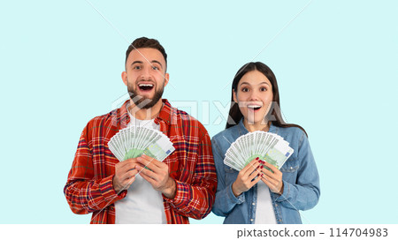 A young man and woman stand against a blue background, expressing surprise and happiness while holding a fan of cash 114704983