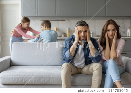 A european family exhibits a mix of love, affection, and stress as overwhelmed parents sit on the couch while their children play in the background, highlighting complex family relationships 114705045