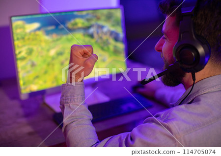 A gamer guy with a fist clenched in a moment of triumph or intense gameplay, engaged with the computer screen 114705074