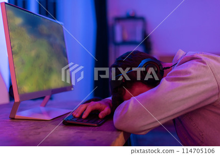 A tired man with headphones appears to be sleeping or frustrated at a desk with a computer screen glowing in a dimly lit room 114705106