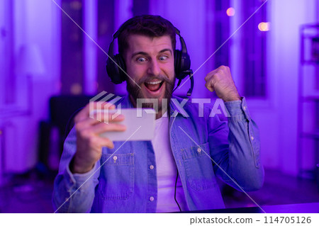 An overjoyed guy gamer uses a gaming headset while playing on his smartphone, celebrating a moment in a neon-lit room 114705126