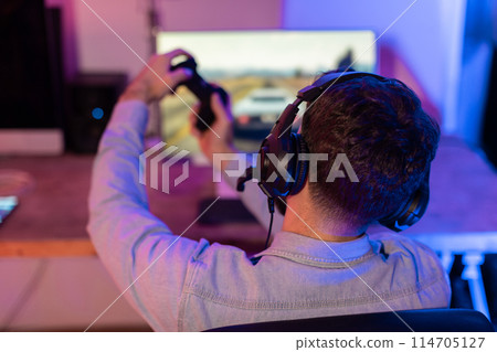 Gamer man with headphones raises arms in the air in victory or excitement, behind a computer monitor, back view Gamer man with headphones raises arms in the air in victory or excitement, behind a computer monitor, back view 114705127
