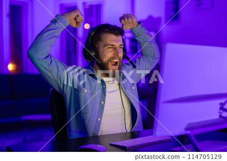 Gamer guy celebrates a victory with enthusiastic cheers, surrounded by blue light in a gaming setup 114705129