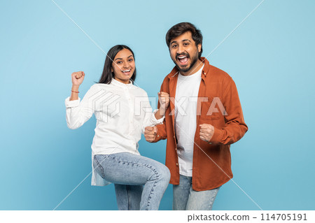 An exuberant Indian couple with fists raised celebrating victory or success together with joy on blue 114705191