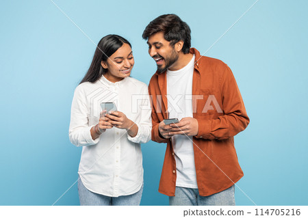 A happy Indian man and woman smile as they share content on a smartphone, conveying connection on blue 114705216