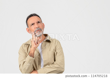 Thoughtful senior man in casual attire looks up with finger on cheek, signifying deep in thought or making a decision, isolated on white background Thoughtful senior man in casual attire looks up with finger on cheek, signifying deep in thought or making a decision, isolated on white background 114705218