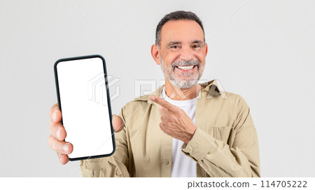 A cheerful senior man with stubble in a casual shirt holding up a smartphone with a blank white screen, pointing at it with his other hand 114705222