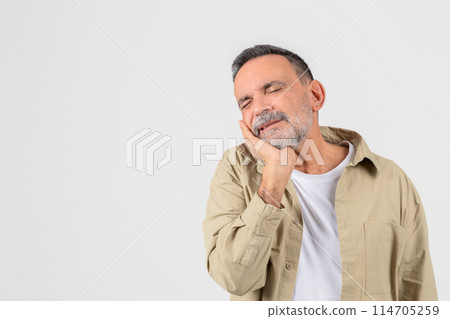 A senior man holds his cheek, expressing discomfort, possibly due to a dental issue or toothache, against a clean white background A senior man holds his cheek, expressing discomfort, possibly due to a dental issue or toothache, against a clean white background 114705259